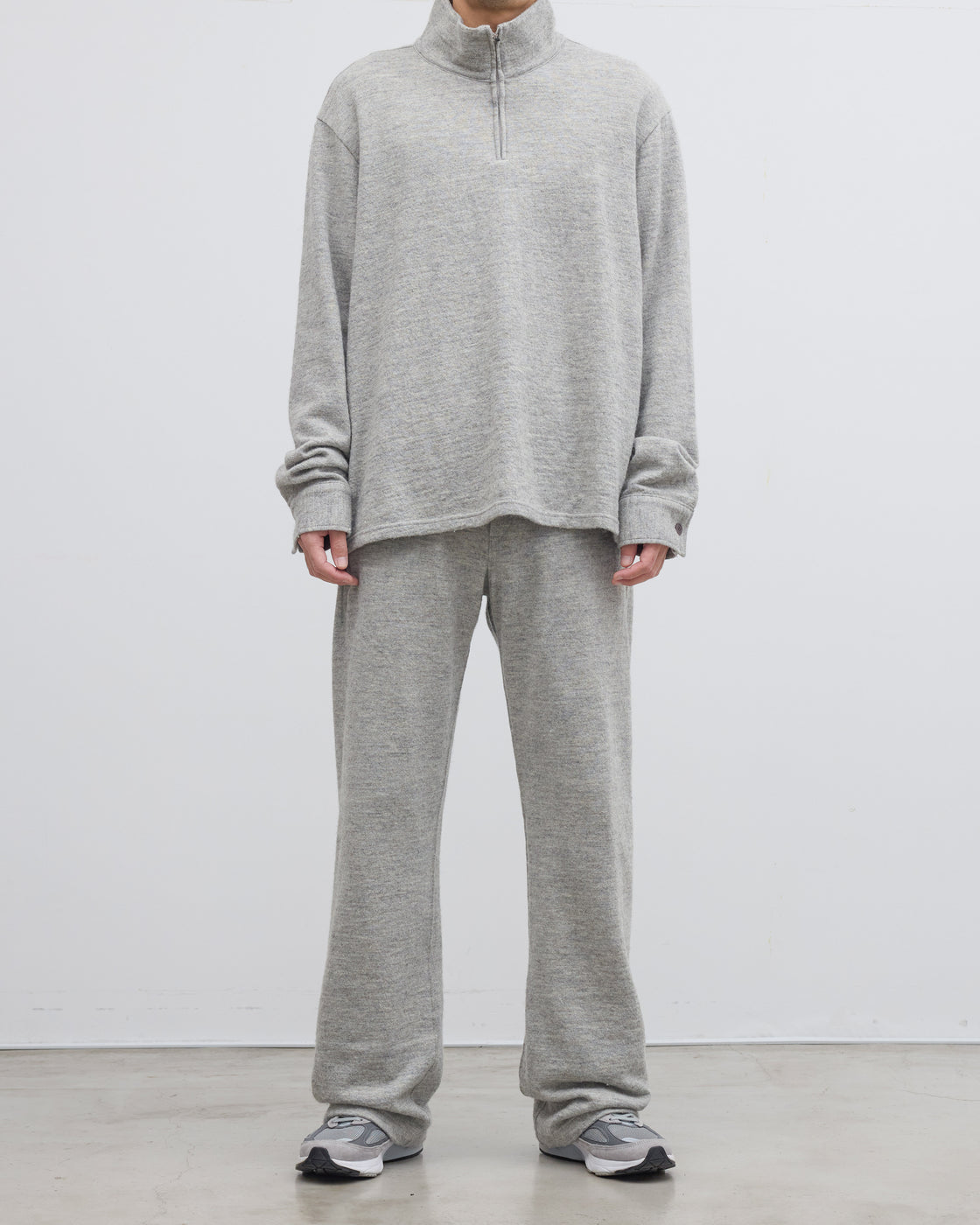 グレーのメランジ糸ハーフジップニットプルオーバーを着用したモデルの後ろ姿、シェットランドウールとコットンの撚り合わせによる独特な風合いと尾州産ニットの高い品質が感じられる一枚